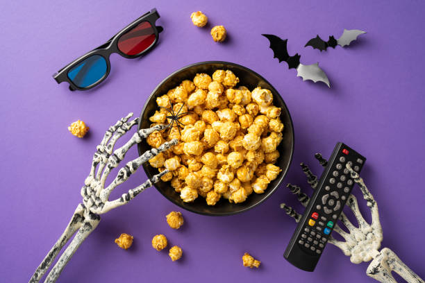 istock Skeleton hands grabbing popcorn with a remote on a purple background, highlighting a spooky Halloween movie night concept 2166202900