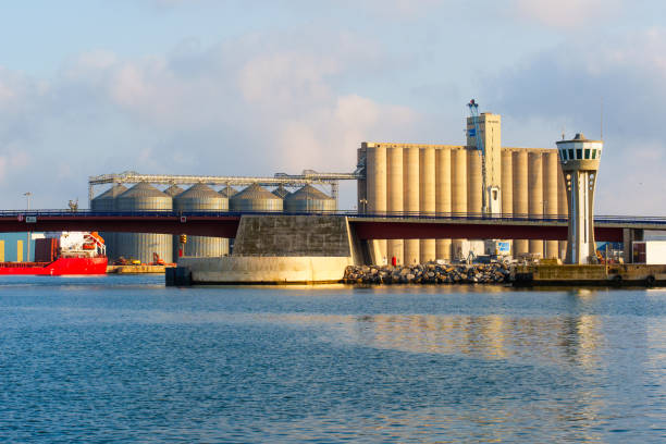 Tarragona, Spain - July 15, 2024: View of large silos and watchtower at Tarragona harbor, surrounded by calm water and a partly cloudy sky, with cargo ship moored nearby.