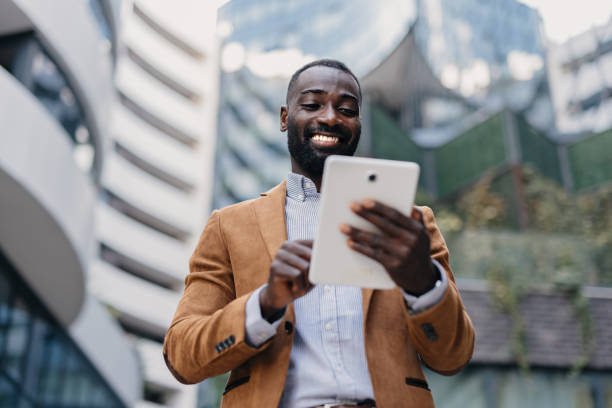 Man smiling while interacting with a tablet device A professional man smiles as he interacts with a tablet device outdoors, demonstrating the joy and convenience of using technology in a business-oriented setting. african american architecture stock pictures, royalty-free photos & images