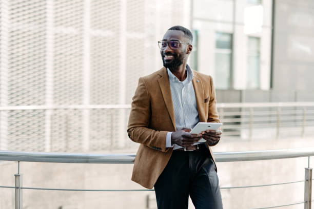 Man leaning on railing with a tablet outdoors A stylish man in a brown jacket and glasses leans on a railing outdoors, looking thoughtful while holding a tablet, representing modern urban sophistication and connectivity. african american architecture stock pictures, royalty-free photos & images
