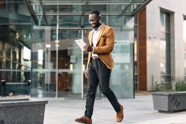 Man walking with tablet by modern office building A professional man in a smart brown jacket walks by a modern office building while holding a tablet, showing the bustling life of contemporary business and technology. african american architecture stock pictures, royalty-free photos & images