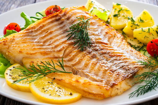 istock Fried cod fillet, boiled potatoes, fresh vegetables and lemon slices served on white plate on wooden table 2166220416