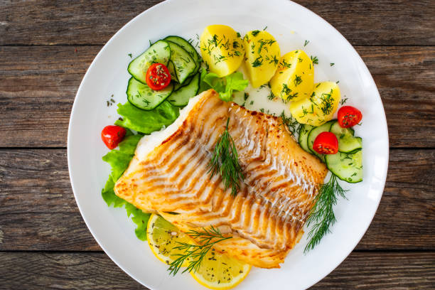 istock Fried cod fillet, boiled potatoes, fresh vegetables and lemon slices served on white plate on wooden table 2166220477