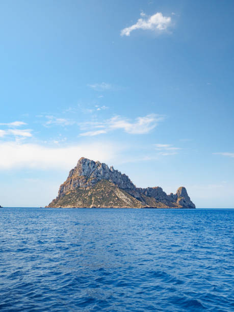Daytime photo of Es Vedra island, off the coast of Ibiza, taken from the sea, with blue skies and and a few clouds visible on the horizon