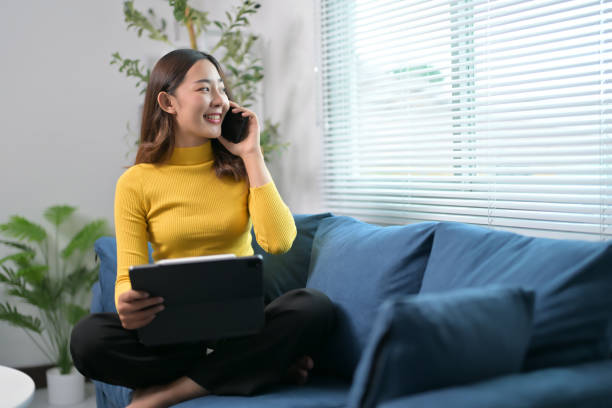 istock Young Woman in Yellow Sweater Talking on Phone While Using Tablet on Blue Sofa at Home 2166235820