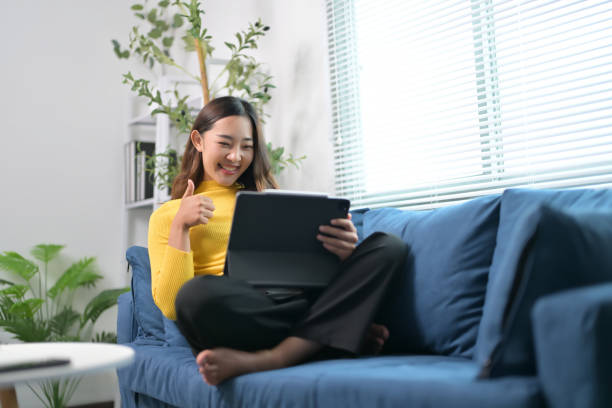 istock Young Woman Smiling and Giving Thumbs Up While Using Tablet on Comfortable Blue Sofa in Modern Living Room 2166247242
