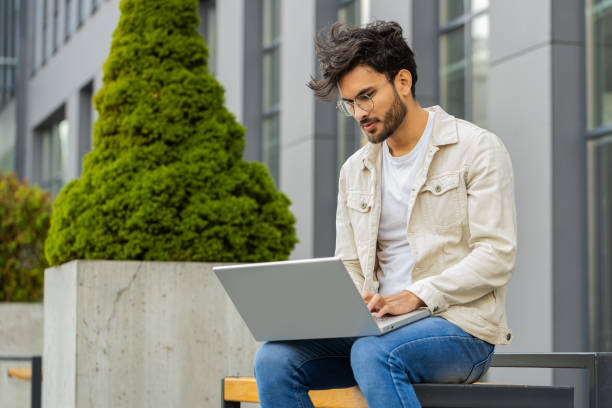 istock Indian man freelancer working on laptop computer sends messages reading email outdoors on bench 2166247403