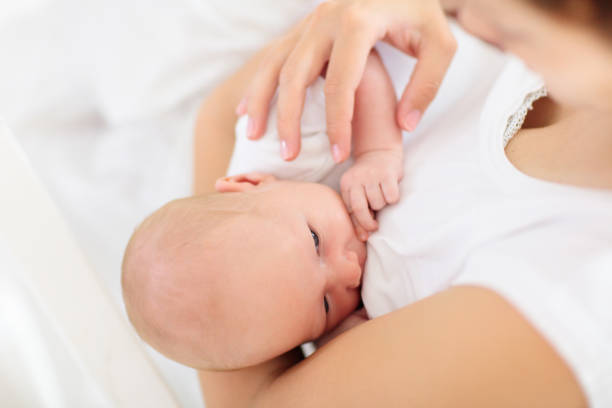 Young mother with newborn baby. Mom nursing little baby boy. Family at home. Infant care. Breastfeeding for kids health. White bedroom for little child.