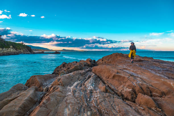 A female traveler is walking on top of the cliff away from the sea at sunset time with dramatic clouds and sunset hue at the background.