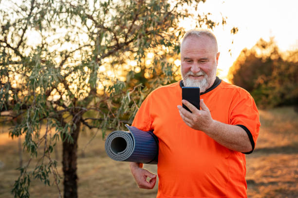 Sporty mature man using phone, holding exercise mat outdoors in the park stock photo