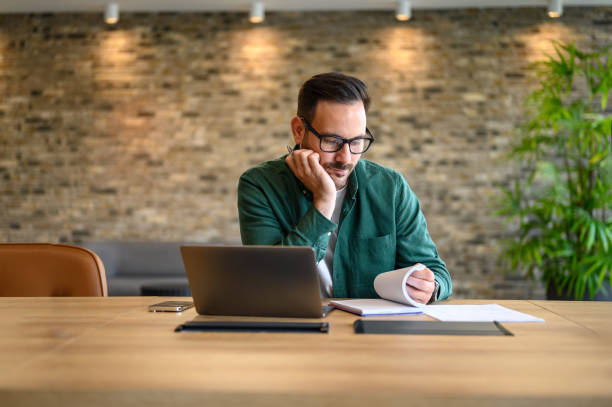 istock Male project manager in eyeglasses reading notes while working online over laptop at desk in office 2166286732
