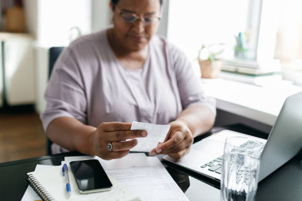 Selective focus on black female hands holding utility bill or paycheck sitting in front of laptop with smartphone on table near window at kitchen, puzzled with high prices due to inflation