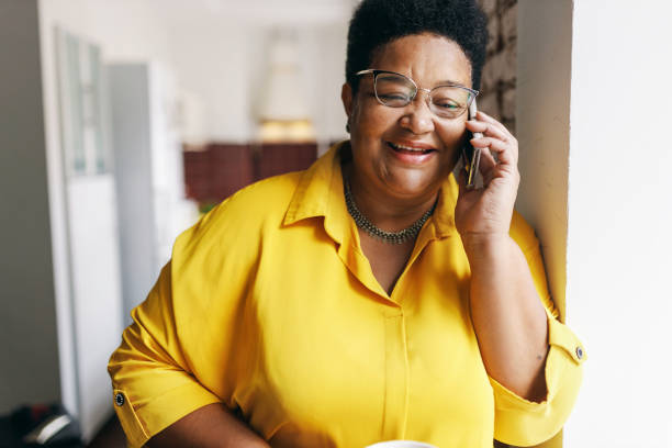 happy smiling senior black plus size female in yellow shirt and glasses having nice conversation on phone with her best friend, smiling and laughing at jokes, sharing news and rumors - plussize model fotos stockfoto's en -beelden