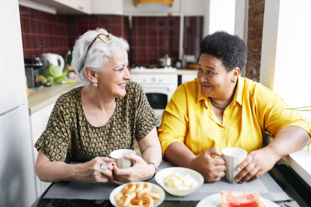 istock Two aged ladies of diverse ethnicity sitting at kitchen table drinking coffee, sharing gossips and rumors, discussing news, looking at each other with laughing faces, enjoying spending time together 2166298312