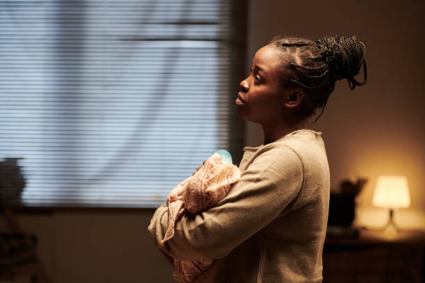 Side view of young vulnerable and lonely African American woman with newborn baby in hands standing in front of camera at home