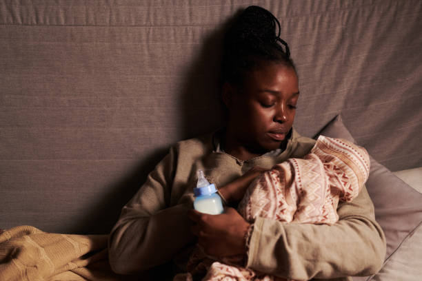 African American single mother looking at newborn baby wrapped in cloth while sitting on couch in front of camera and lulling infant