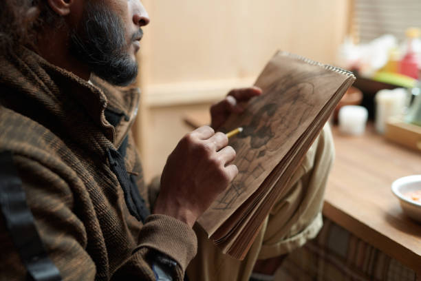 Man focusing on detailed sketching in spiral notebook while sitting in cozy indoor setting with table and food nearby creating artistic drawing