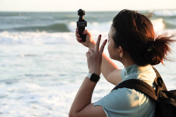 istock Young asian woman wearing backpack is taking a selfie at the beach using pocket gimbal camera with peace sign hand gesture. Concept for solo traveling. 2166331010