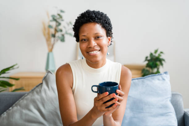 istock Young black woman smiling and holding a mug while sitting on a sofa 2166353114