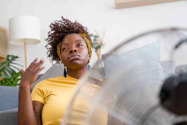 istock Young woman sweating on sofa at home using fan feeling hot 2166355860