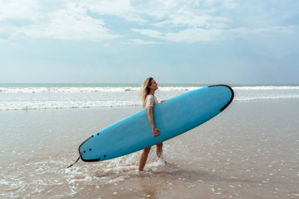 istock Cheerful woman carries a blue surfboard while walking on the beach 2166358476