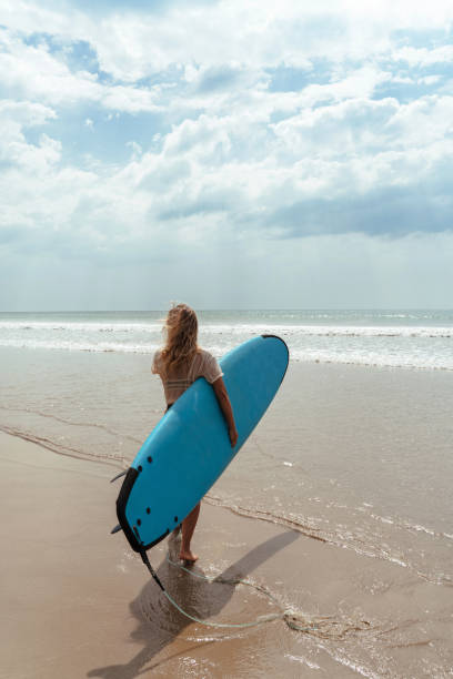 istock Cheerful woman carries a blue surfboard while walking on the beach 2166358582