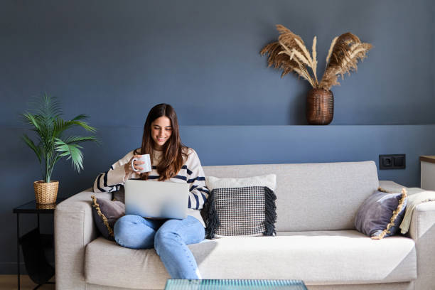 istock Beautiful woman sitting on a comfortable sofa using a laptop and drinking coffee in a modern living room 2166365878