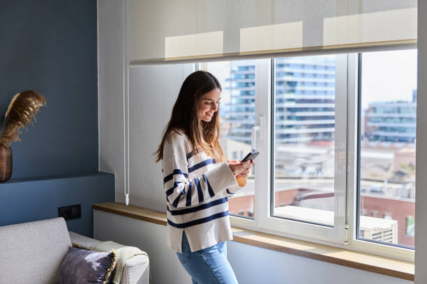 Happy woman is using a smartphone while standing by the window in a modern living room