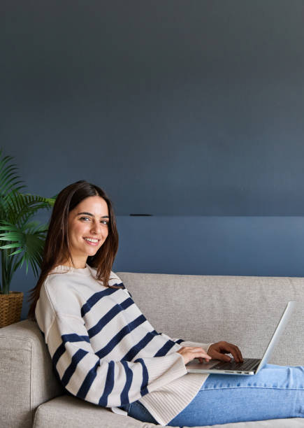 istock Portrait of a beautiful woman smiling at the camera while using a laptop computer on her sofa at home 2166366035