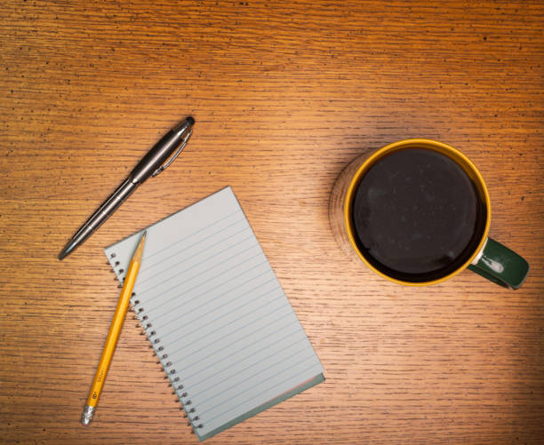 Coffee in gold and green trimmed cup sitting on a rough wood plank table next to plank spiral note pad and pencil