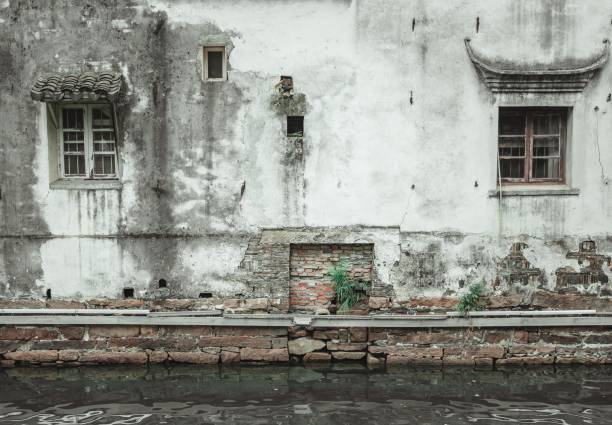 A weathered building facade with windows and a brick wall, reflected in calm water.