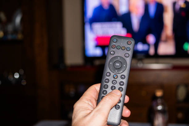 istock Hand Holding Remote Control with Blurred Television in Background. A persons leisure time, controlling media consumption with technology, captured using direct flash photography technique ugc 2166404821
