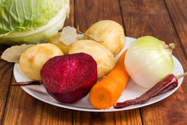 Set of the raw fresh peeled vegetables, white cabbage and dried spices, as ingredients for red beet borscht preparation on a rustic table, side view in selective focus