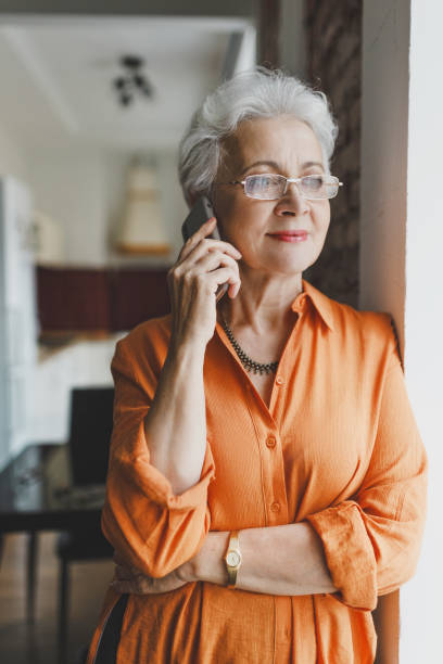 Pretty mature lady with short haircut waiting online while calling to customer support service, standing in kitchen leaning on wall, looking through window, putting hand on her belly