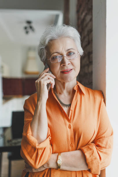 istock Pretty mature lady with short haircut waiting online while calling to customer support service, standing in kitchen leaning on wall, looking through window, putting hand on her belly 2166427542