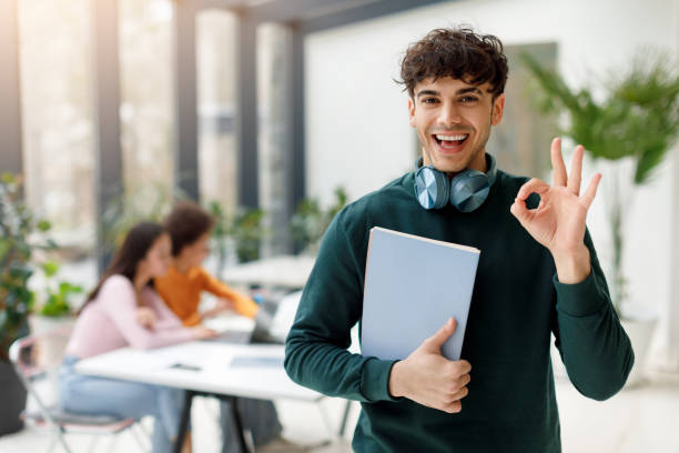 Cheerful male student with OK gesture holding book