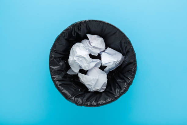 Crumpled white paper in black opened plastic garbage bag on light blue floor background. Pastel color. Closeup. Top down view.