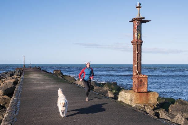 Castlerock, UK - March 5th, 2022: Woman exercising with white dog along the pier at the Barmouth where the River Bann enters the sea.