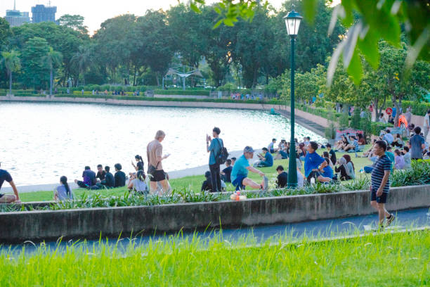 Young thai people are relaxing in Benjakitti Park at sunset. Park is located in Bangkok at Ratchadaphisek Rd, Khlong Toe. People are sitting and relaxing on grass between pedestrian walkway around the  lake. Many of them are using mobile phones. Other  people are walking in sports fashion or jogging around lake on walkway.  A tall blond young white man is walking with mobile phone in hand on walkway