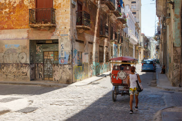 Havana, Cuba - July 30, 2024: A Cuban woman walks on a cobblestone city street in Old Havana. Run-down buildings and a bici-taxi on the scene.