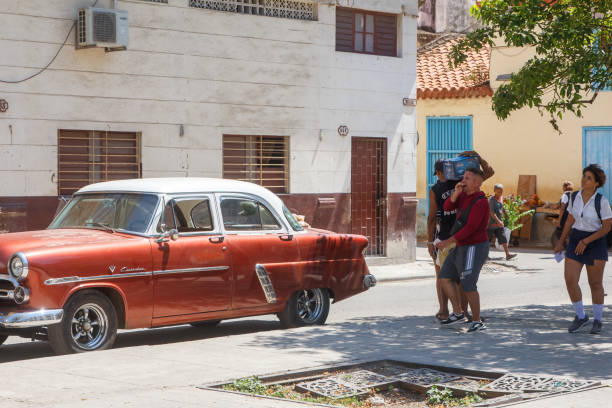 Havana, Cuba - July 30, 2024: A group of Cuban people walk by a vintage American car parked in Old Havana. A part of the sidewalk is broken.