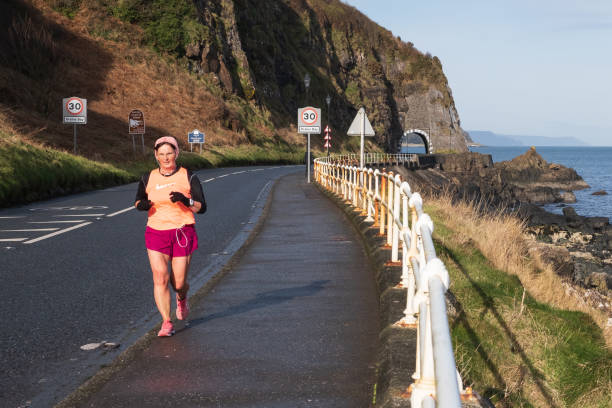 Drains Bay, UK - February 11th, 2022: Female lone runner taking exercise just outside coastal town with the Black Arch in the background and views of the shoreline and ocean.