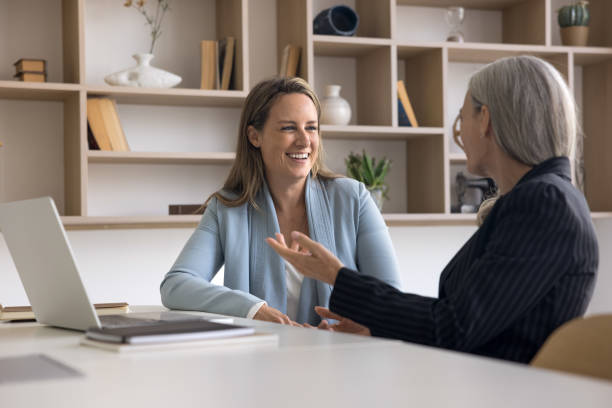 Two positive younger and elder businesswomen talking at workplace