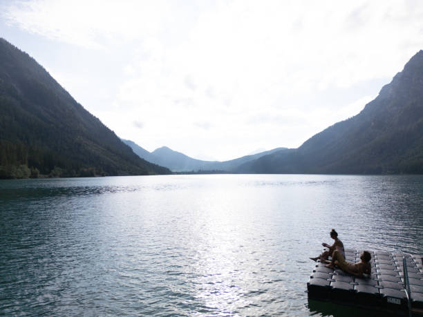 A couple lies down on a floating dock, enjoying a peaceful moment by a scenic mountain lake under a partly cloudy sky. The serene setting reflects tranquility and connection with nature.