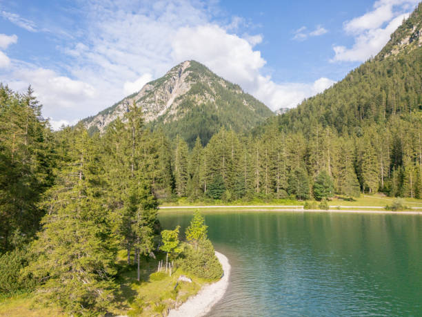 A serene mountain lake with a forested shoreline, surrounded by towering peaks under a bright, partly cloudy sky. The scene captures the natural beauty and tranquility of the wilderness.