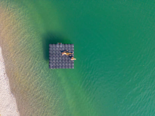 Aerial View of Couple Relaxing on Floating Dock in Clear Lake An aerial shot of a couple on a floating dock in a clear, calm lake. The top-down perspective emphasizes the tranquil water and the simplicity of outdoor relaxation. jetty aerial stock pictures, royalty-free photos & images