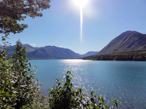 The sun is casting a radiant glow over a crystal-clear mountain lake in Alaska. The towering peaks are reflected in the still waters, creating a serene and picturesque scene.