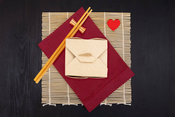 Wok box for asian food with chopsticks. Black wooden background, yellow bamboo mat. Red cloth napkin. Small red heart.