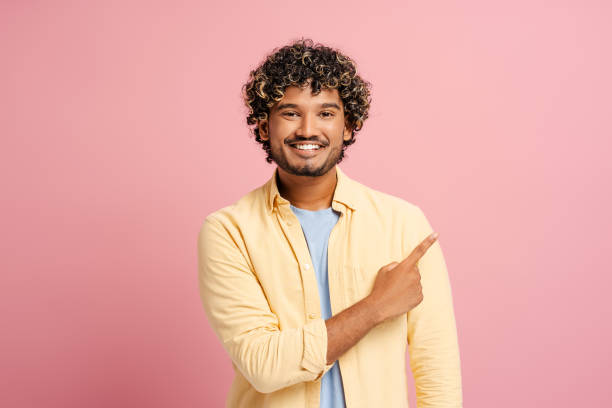 istock Cheerful Indian young man pointing with finger aside on pink background, advertisement concept 2166513451