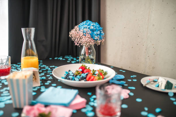 A gender reveal party setting indoors, showcasing a table with blue confetti, indicating a baby boy. Pink and blue napkins, a fruit plate, and drinks complement the celebratory arrangement.